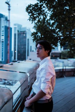 Beyaz tişörtlü, gözlüklü, siyah saçlı adam şehrin çatısında dikiliyor. Şehrin rüzgârını ve gece manzarasını seyrediyor..