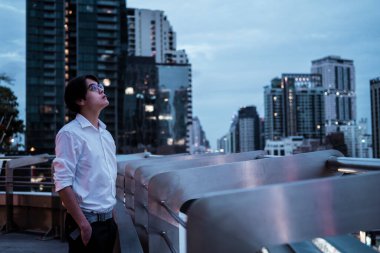 Beyaz tişörtlü, gözlüklü, siyah saçlı adam şehrin çatısında dikiliyor. Şehrin rüzgârını ve gece manzarasını seyrediyor..