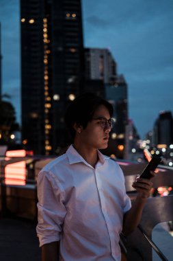 Beyaz tişörtlü ve gözlüklü siyah adam çatıda duruyor. Kablolu yaylı barikatla. Akıllı telefonunu bina, yol ve arabaları kullanıyor..