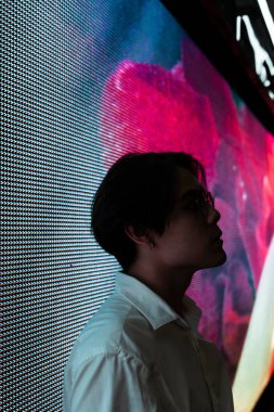 Gözlüklü siyah saçlı adamın, reklam için LED reklam panosunun önünde durduğu ve geceleri şehirdeki alışveriş merkezinin reklamını yaptığı bir fotoğraf..