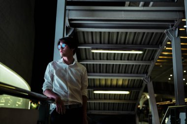 Beyaz tişörtlü ve gözlüklü siyah saçlı adam Skytrain istasyonunun yürüyen merdiveninde duruyor ve kolunu tırabzana dayıyor..