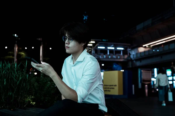 Black hair guy sitting on the footpath next to the skytrain station and use smartphone.