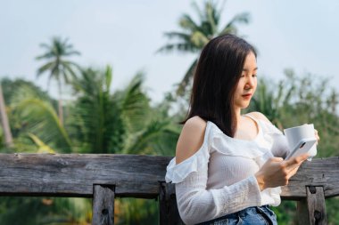 Beyaz tişörtlü mutlu Asyalı kadın elinde bir fincan kahve ve tatilde arka planda tatil yeri ya da çiftlik ile akıllı telefon oyunu oynuyor..
