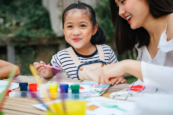 Asyalı abla, küçük kız kardeşine açık havada su rengini boyamasında yardım ediyor..