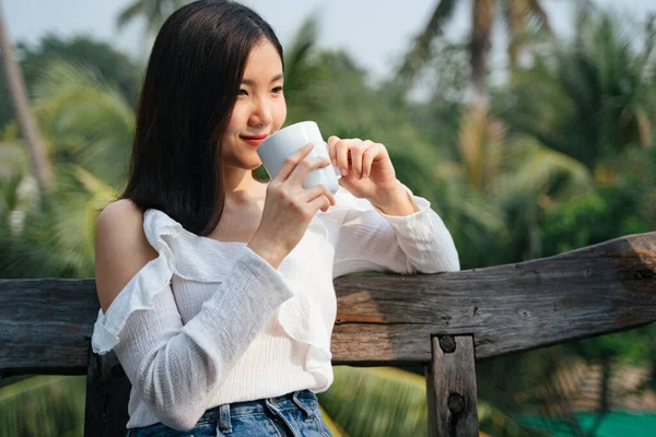 Thai asian woman sitting on wooden bench drinking a cup of coffee at countryside district. Travel in nature. Aroma coffee in a cup. Gorgeous woman drinking coffee from white cup.