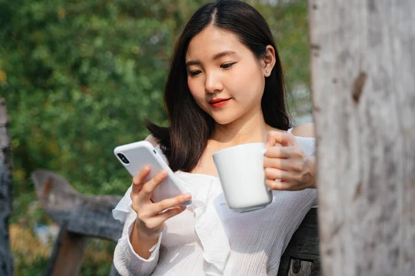 Asian thai woman in white shirt holding a cup of coffee, sitting on wooden bench, using smartphone to connect with friend.