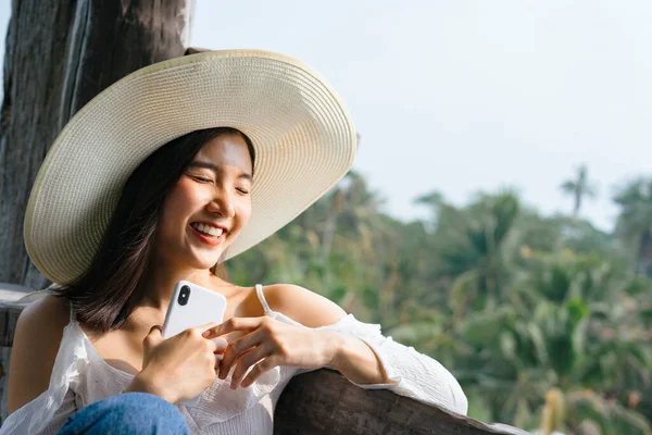 陽気なアジアの女性が帽子をかぶってスマートフォンを持ってバルコニー田舎に座って — ストック写真