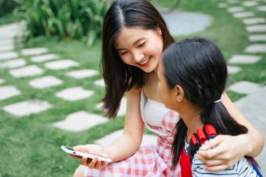Neşeli iki Asyalı kız kardeş bahçede oturup akıllı telefon oynamaktan zevk alıyor..
