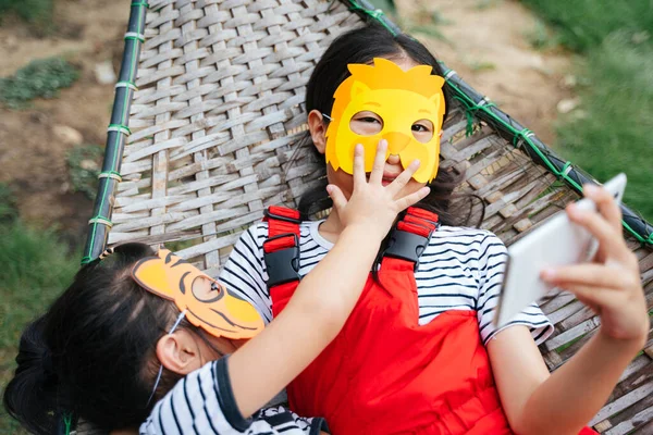 Cheerful asian little girls wearing cartoon animal mask laying on a cot playing smartphone. Little sister trying to steal mobile phone from big sister.