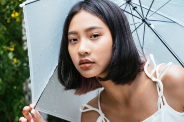 Bahçe parkında beyaz şemsiye tutan genç Taylandlı kadına yakın durun..