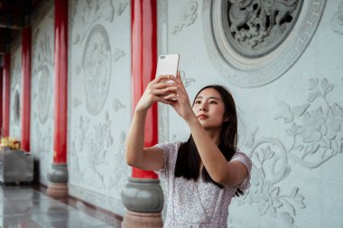 Geleneksel Çin tapınağında akıllı telefonuyla selfie çeken sevimli Asyalı genç bir kadın. .