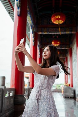 Geleneksel Çin tapınağında akıllı telefonuyla selfie çeken sevimli Asyalı genç bir kadın. .