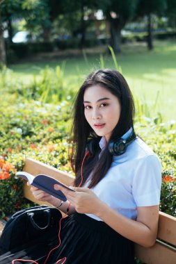 Uzun siyah saçlı üniversiteli kız tahta bankta oturuyor, kitabı tutuyor ve kameraya bakıyor..