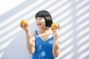 Neşeli, mutlu, güzel, kısa saçlı, mavi kolsuz tişörtlü Asyalı kadın elinde iki portakal, elinde tropik meyve, beyaz arka planda..