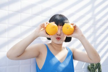Komik, güzel, kısa saçlı, mavi kolsuz tişörtlü Asyalı kadın elinde portakal tutarken gözlük gibi davranıyor..