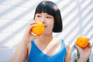 Sağlıklı, kısa saçlı, mavi kolsuz tişörtlü Asyalı güzel bir kadın tropikal portakal meyvesi yiyor..