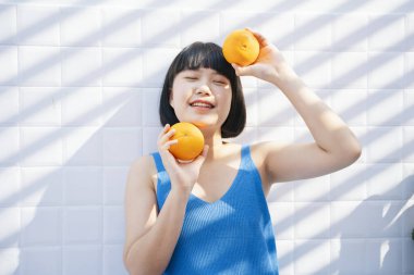 Mavi tişörtlü Asyalı güzel kısa saçlı kadın elinde iki portakal, beyaz arka planda ise tropik meyve tutuyor..