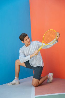 Mavi turuncu arka planda tenis raketi tutan sıradan kostümlü çekici Asyalı bir genç. Komik poz. Tenis oynuyordum. Yerde otururken topa vurmaya hazır..