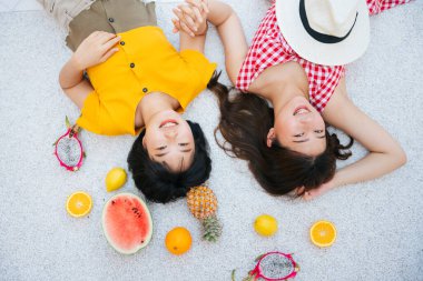 Yaz tatilinde yerde yatan Asyalı iki yakın arkadaş. Tropik meyve. Üst Manzara. Sarı tişörtlü ve kırmızı elbiseli Asyalı kadın. Seyahat tatili. Hafta sonu tatili.