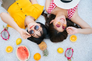 Yerde tropik meyvelerle yatan iki Asyalı arkadaş. Sağlıklı meyveler. Üst Manzara. Yaz tatili.