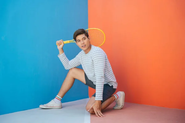 Attractive young asian man in casual costume holding tennis racket over blue orange background. Funny posing. Playing tennis. Cool posing with the racket gesture.