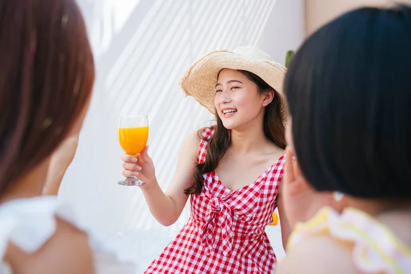 Beautiful young asian woman wearing hat and red dress drinking orange juice with friends in summer party.