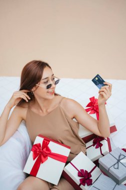 Güneş gözlüğü takan genç ve güzel bir kadın beyaz koltukta oturuyor. Bir sürü hediye kutusu ve kredi kartı var. Tatilde online alışveriş. Bej krem arka planda izole edilmiş..