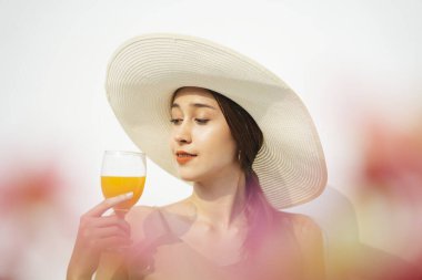 Çekici, güzel esmer kadın beyaz arka planda izole edilmiş cam bardakta büyük bir şapka ve portakal suyu giyiyor..