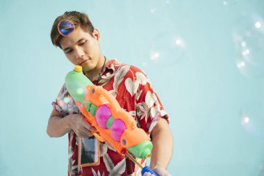 Yakışıklı Taylandlı adam elinde su tabancası tutan çiçek bezi giymiş. Yumuşak mavi arka planda izole edilmiş. Tayland 'da Songkran Festivali.