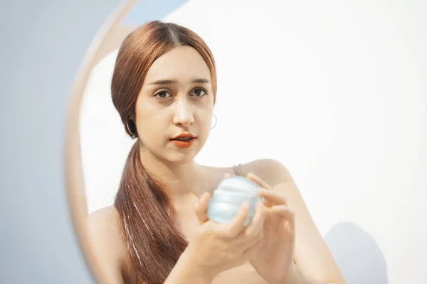 Attractive beautiful brunette woman holding cosmetic jar applying make up in front of the mirror.