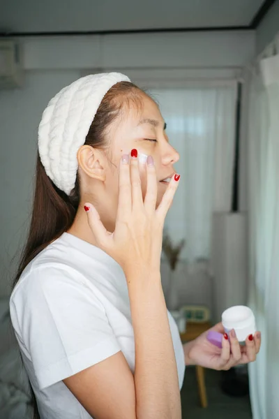 Woman with nail painting in white t-shirt and headband apply facial cream on her face.