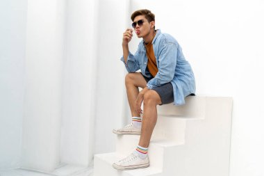 Genç Asyalı yakışıklı Taylandlı adam kot ceket giyiyor ve güneş gözlüğü takıyor beyaz merdivende oturuyor lolipop şekeri yalıyor..