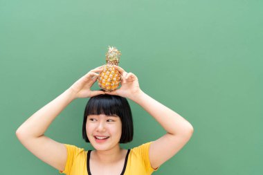 Bob saç stili genç Taylandlı kadın kafasında ananas tutuyor. Yeşil arkaplan üzerinde izole edilmiş.