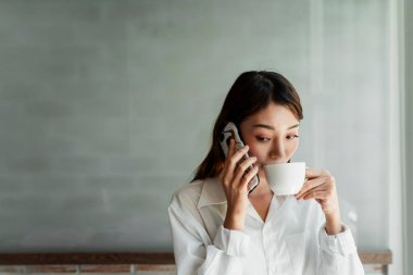 Uzun saçlı, beyaz gömlekli kadın iç mimar müşterisinin telefonuna cevap vermesini beklerken sıcak kahve içiyor..