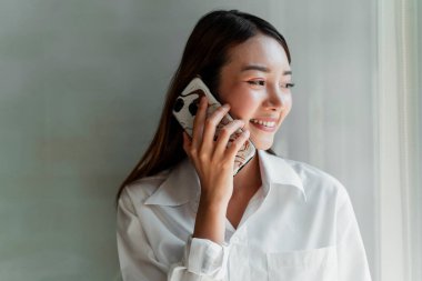 Uzun saçlı, beyaz tişörtlü kadın pencerenin yanında duruyor. Telefonda biriyle konuşuyor..