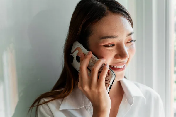 Long hair designer woman in white shirt standing at the wall next to the window talkig to someone on the phone.