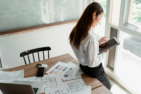 Long hair designer woman in white shirt sitting on the table in the office and manage her to do list on tablet.