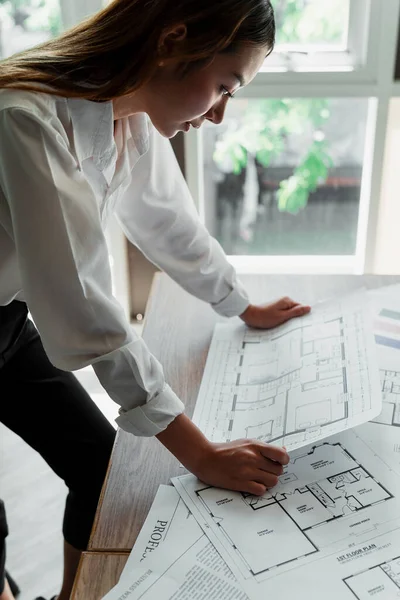 Long hair designer woman in white shirt standing at her wooden table look at the building plan in her hands.