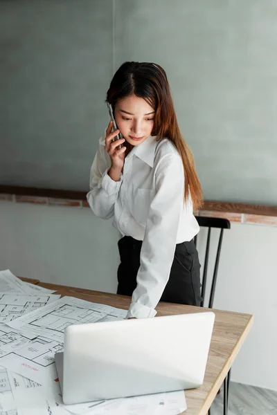Long hair designer woman in white shirt standing at her wooden table look at the building plan and conpare with the plan in her laptop, talking to her team on the phone.