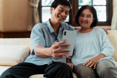 Bir çift Asyalı yaşlı insan akıllı telefonuyla selfie çekiyor, gülümsüyor, kanepede oturuyor..
