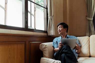 Yalnız Asyalı yaşlı adam kanepede oturup tablet kullanıyor..