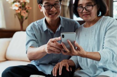 Oturma odasında kanepede oturmuş akıllı telefon oynayan sağlıklı yaşlı bir çift. Üst düzey aileler akıllı telefonu birlikte kullanmaktan hoşlanırlar..