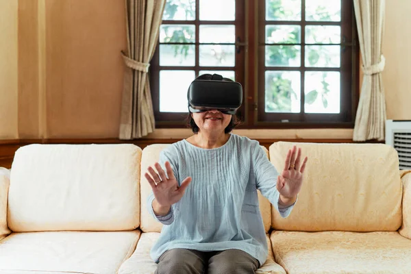 technology, augmented reality, entertainment and people concept - senior woman with virtual headset or 3d glasses playing videogame at home on a couch.