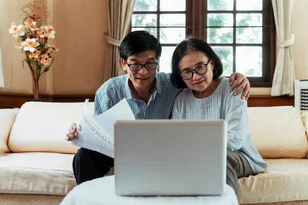 Asian elderly couple sitting on a couch, looking laptop, video calling with their kids. Internet is useful for old people who stay at home.