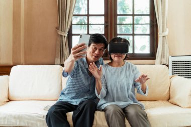 Yaşlı Asyalı çift kanepede oturup VR gözlüğü takıyor ve akıllı telefonuyla selfie çekiyor..