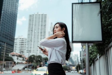 Beyaz tişörtlü Asyalı iş kadını sokakta akıllı telefon kullanıyor ve bir arabanın onu almasını bekliyor. Boşluğu kopyala.