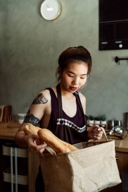 Asyalı bir kadın bir çift Fransız ekmeği alıyor ve onları kağıt poşete koyuyor..