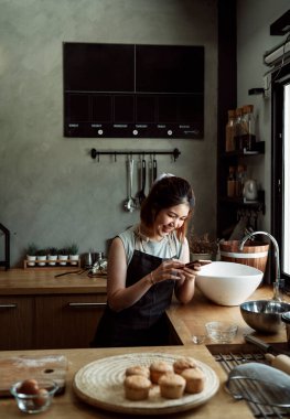 Neşeli Asyalı kadın pencerenin yanındaki mutfakta akıllı telefon kullanıyor..