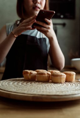 Asyalı fırıncı kadın, sosyal medya için akıllı telefonuyla mutfaktaki ahşap tabakta taze kekin fotoğrafını çekiyor..