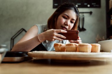 Asyalı fırıncı kadın, sosyal medya için akıllı telefonuyla mutfaktaki ahşap tabakta taze kekin fotoğrafını çekiyor..